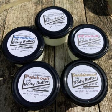 5 clear glass jars with black lids containing 5 different scented body butters: baby powder, lavender, patchouli, rose, sandalwood